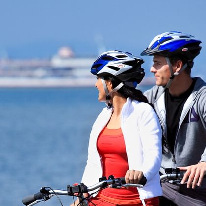 Zwei Radfahrer mit Helmen stehen mit ihren Rädern am Bodensee und blicken auf eine Fähre auf dem blauen Wasser mit Bergen im Hintergrund.