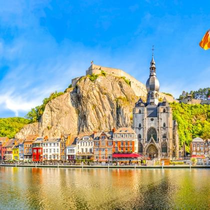 Stadtansicht von Dinant mit bunten Häusern an der Maas, gotischer Kirche und Zitadelle auf Felsenklippe unter blauem Himmel mit belgischer Flagge.