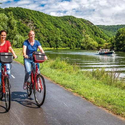 Zwei Frauen radeln auf asphaltiertem Weg entlang eines Flusses in Luxemburg mit bewaldeten Hügeln und Ausflugsboot im Hintergrund.