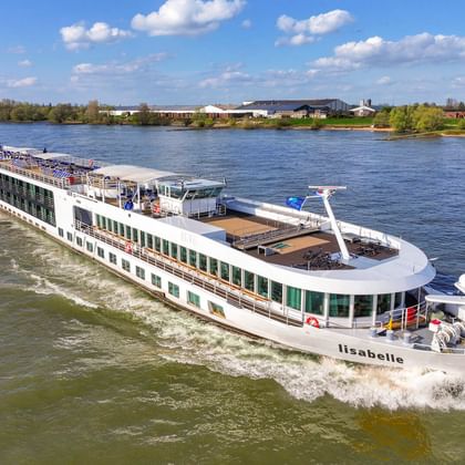 Weißes Flusskreuzfahrtschiff MS Lisabelle fährt auf einem breiten Fluss unter blauem Himmel mit Wolken. Das moderne Schiff hat mehrere Decks und Panoramafenster.