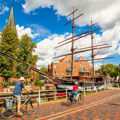 Zwei Radfahrer auf einem Backsteinweg neben einem historischen Segelschiff in Papenburg. Kirchturm und orangefarbene Backsteingebäude unter blauem Himmel.