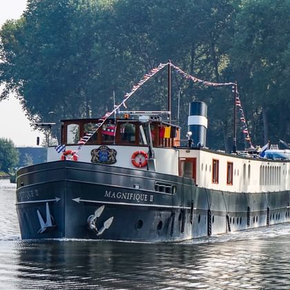 Passagierschiff Magnifique II mit dunklem Rumpf und weißer Kabine auf ruhigem Kanal, gesäumt von dichten grünen Bäumen unter klarem Himmel.