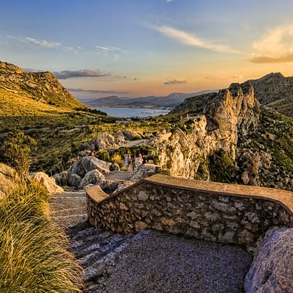 Steinerner Aussichtspunkt mit Felsformationen in Mallorcas Bergen bei Sonnenuntergang, mit Blick auf Küstenlandschaft und Meer unter goldenem Himmel.
