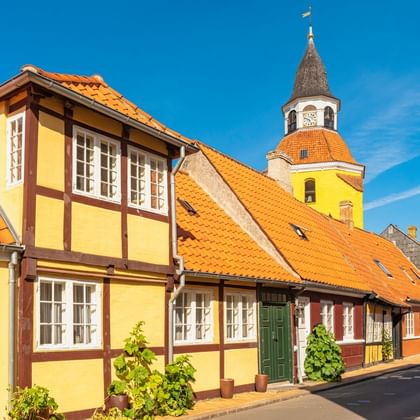 Historische Straße in Faaborg mit gelben Fachwerkhäusern und orangen Ziegeldächern. Kirchenglockenturm mit Uhr im Hintergrund unter blauem Himmel.