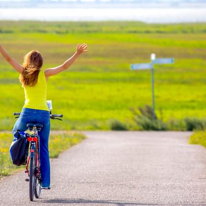 Frau auf Fahrrad mit freudig erhobenen Armen auf Weg durch grüne Wiesen in Ahrenshoop. Ein weißer Wegweiser steht neben dem Weg.