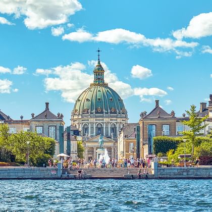 Blick über das Wasser zur Frederikskirche mit ihrer markanten blaugrünen Kupferkuppel, umgeben von historischen Gebäuden und Bäumen unter blauem Himmel.