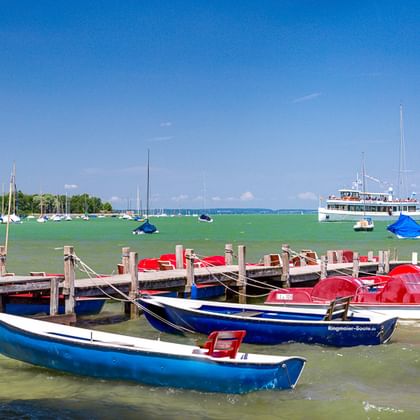 Blaue und rote Tretboote an einem Holzsteg an einem bayerischen See. Grünes Wasser, Segelboote und ein weißes Ausflugsschiff im Hintergrund.