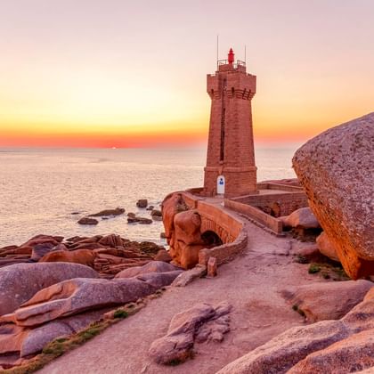 Rosa Granitleuchtturm von Ploumanac'h bei Perros-Guirec, umgeben von großen rosa Felsen bei Sonnenuntergang mit Meer und orangefarbenem Himmel.