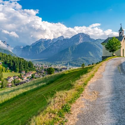 Kleine Kapelle mit Glockenturm auf grünem Hügel bei Toblach, Südtirol. Ein Schotterweg führt zur Kapelle mit Berggipfeln im Hintergrund.