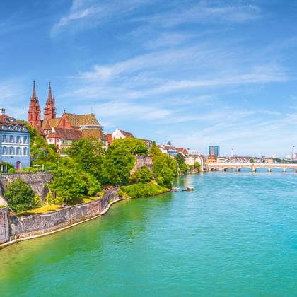 Türkisfarbener Rhein fließt durch Basel mit historischen Gebäuden, roten Kirchtürmen und Steinmauer unter blauem Himmel.