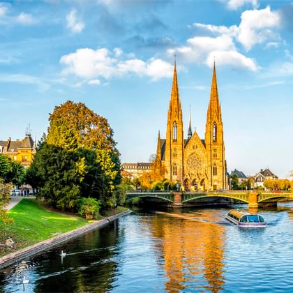 Gothic church with twin spires in Strasbourg beside a canal with a boat. Green park and bridge in foreground under blue sky with clouds.