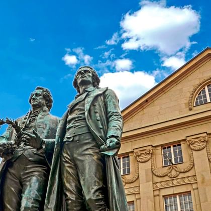 Bronzenes Goethe-Schiller-Denkmal in Weimar mit zwei stehenden Figuren vor blauem Himmel und klassizistischem Theatergebäude mit Rundbogenfenster.