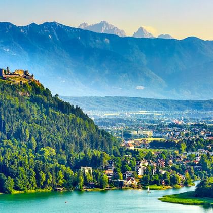Türkisfarbener See mit bewaldeten Hügeln, Burg auf Hügel, Stadt im Tal und Bergkette unter blauem Himmel am Alpe Adria Radweg.