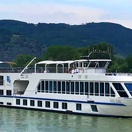 Weißes Flusskreuzfahrtschiff MS SE-Manon mit blauem Streifendesign fährt auf ruhigem Wasser vor grünen Bergen.
