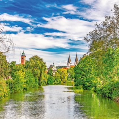 Blick auf die Altmühl in Eichstätt mit üppig grünen Bäumen am Ufer und historischen Kirchtürmen im Hintergrund unter blauem Himmel.