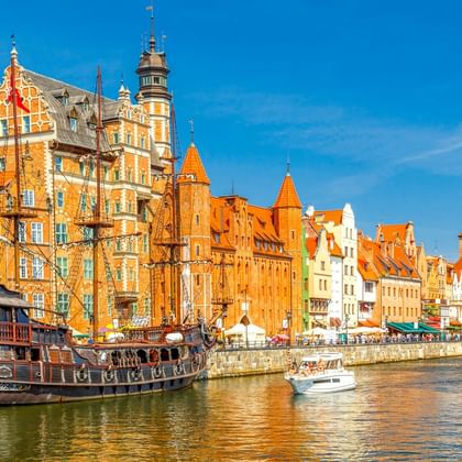 Bunte historische Gebäude entlang der Danziger Uferpromenade mit Segelschiff-Nachbau und mittelalterlichem Kranturm unter blauem Himmel.