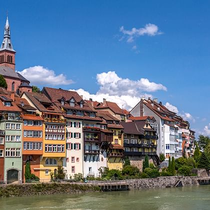 Historische bunte Häuser am Rhein mit Kirchturm auf dem Hügel. Blauer Himmel mit weißen Wolken über der Flussstadt.