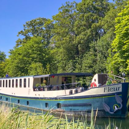 Flusskreuzfahrtschiff MS Fleur an einem von Bäumen gesäumten Kanal in Frankreich. Das weiß-blaue Schiff hat ein offenes Deck mit Passagieren.