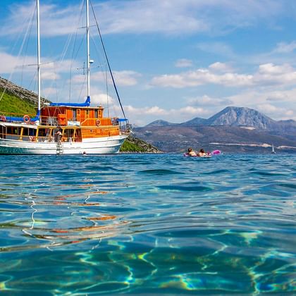 MS Mirabela, ein hölzernes Segelschiff mit zwei Masten, ankert in klarem türkisfarbenem Wasser. Berge und grüner Hang im Hintergrund.