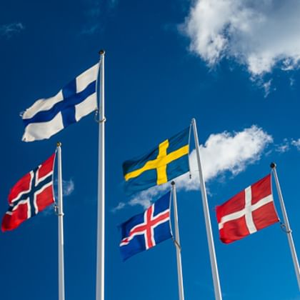 Fünf skandinavische Flaggen wehen im Wind: norwegische, finnische, isländische, schwedische und dänische Flagge vor blauem Himmel mit weißen Wolken.