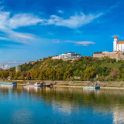 Bratislavaer Burg auf einem grünen Hügel mit Blick auf die Donau mit Kreuzfahrtschiffen unter blauem Himmel mit weißen Wolken.