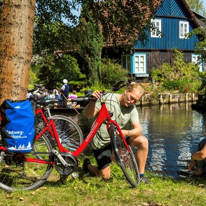 Zwei Radfahrer mit roten Rädern und blauen Taschen rasten an einem Kanal im Spreewald. Ein blaues Fachwerkhaus und ein Boot sind im Hintergrund sichtbar.