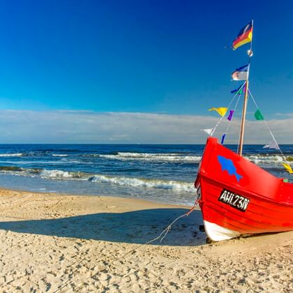 Rotes Fischerboot mit bunten Flaggen am Sandstrand in Ahlbeck, Usedom. Blaue Ostsee mit sanften Wellen unter klarem Himmel.