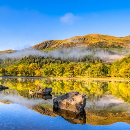 Ruhiger See in Highland Perthshire spiegelt Felsen, Bäume und goldene Hügel. Morgennebel schwebt über der bewaldeten Uferlinie unter blauem Himmel.