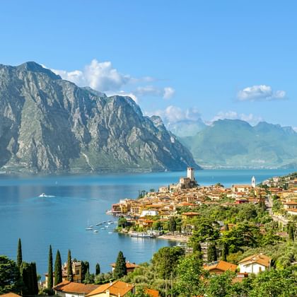 Panoramablick auf den Gardasee mit einer Hanglage-Stadt mit Terrakotta-Dächern, Kirchturm und dramatischen Bergen am blauen Wasser.