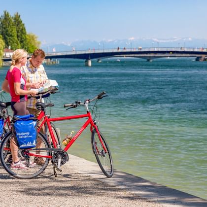 Zwei Radfahrer mit Rädern lesen eine Karte am Seerheinufer in Konstanz. Historischer Turm und Brücke über dem türkisfarbenen Wasser.