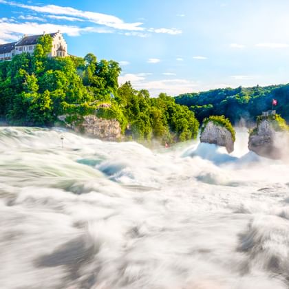 Rheinfall mit kraftvoll strömendem Wasser und Gischt. Schloss Laufen thront auf einer grün bewaldeten Klippe. Eine Schweizer Flagge weht auf einem Felsen.