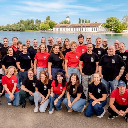 Große Teamgruppe in passenden schwarzen und roten Shirts posiert an einem Fluss mit historischen Gebäuden und Bäumen im Hintergrund.