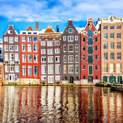 Reihe traditioneller niederländischer Grachtenhäuser in verschiedenen Farben wie Rot, Braun und Orange, die sich im Wasser der Amsterdamer Gracht spiegeln.