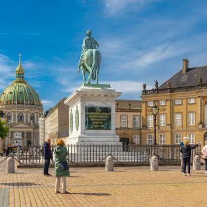 Amalienborg Schlossplatz in Kopenhagen mit Reiterstatue, gelben Palastgebäuden und Kirchenkuppel unter blauem Himmel mit Touristen.