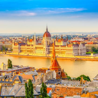 Luftaufnahme des ungarischen Parlamentsgebäudes an der Donau in Budapest, mit historischen Gebäuden und Dächern im Vordergrund.