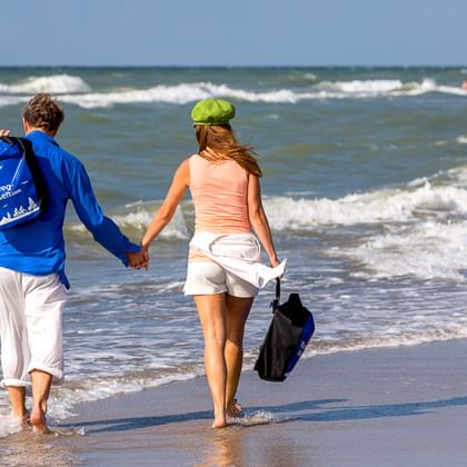 Paar hält Händchen beim Barfußlaufen am Ostseeufer. Mann trägt blaue Radjacke, Frau rosa Top und weiße Shorts.