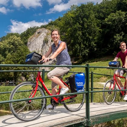 Zwei Radfahrer mit bepackten Rädern überqueren eine Metallbrücke über einen Kanal. Grüne Hügel und Wald im Hintergrund unter blauem Himmel.