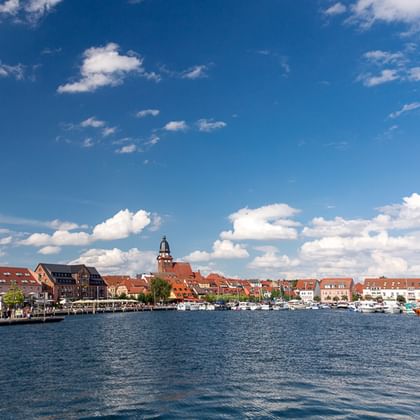 Panoramablick auf eine historische Stadt am Müritz-Ufer mit rotgedeckten Gebäuden, Kirchturm und Booten am Ufer unter blauem Himmel.