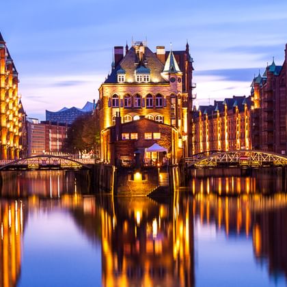Historische rote Backsteingebäude bei Dämmerung beleuchtet, spiegeln sich im Kanalwasser. Brücken verbinden die Gebäude der Speicherstadt.