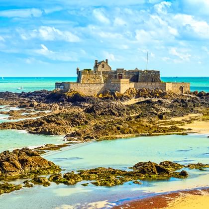 Steinfestung auf felsigem Vorsprung, umgeben von türkisfarbenem Wasser und freiliegenden Felsen bei Ebbe in der Nord-Bretagne unter blauem Himmel.