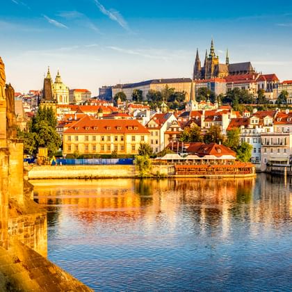 Blick von der Karlsbrücke in Prag mit Statue und Laterne, Moldau, historischen Gebäuden mit roten Dächern und Prager Burg auf dem Hügel.