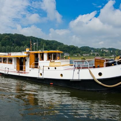 MS Miro, ein weiß-gelbes Flusskreuzfahrtschiff mit schwarzem Rumpf, liegt vor Anker auf ruhigem Wasser mit grünen Hügeln im Hintergrund.