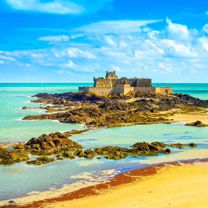 Fort National fortress on rocky island at low tide in Saint-Malo, surrounded by turquoise water, sandy beach, and exposed rocks under blue sky.