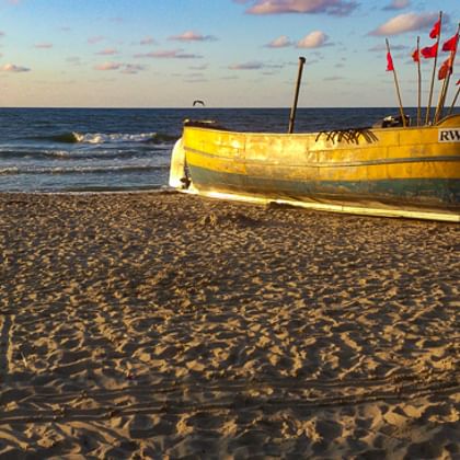 Gelbes Fischerboot mit Kennzeichen RWL-2 und roten Flaggen am Sandstrand bei Sonnenuntergang. Ostseewellen und blauer Himmel mit rosa Wolken im Hintergrund.