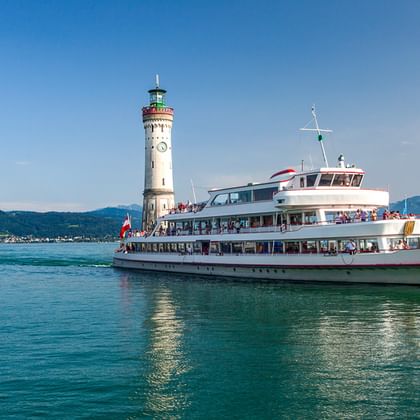 Weiße Personenfähre auf dem Bodensee passiert die Lindauer Hafeneinfahrt mit Leuchtturm und Löwenstatue. Berge im Hintergrund.