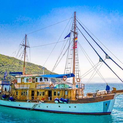 MS Panagiota, ein traditionelles Segelschiff mit türkisfarbenem Rumpf und Holzdeck, vor Anker in ruhigem blauen Wasser bei Parga mit grünen Hügeln.