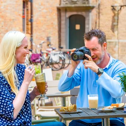 Zwei Radfahrer genießen Getränke an einem Café-Tisch in Middelburg. Mann fotografiert Essen, Frau trinkt. Fahrräder vor historischem Gebäude.