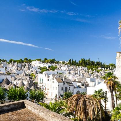 Blick auf traditionelle weiße Trulli-Häuser mit Kegeldächern in Apulien, Italien. Palmen im Vordergrund, blauer Himmel über dem historischen Dorf.