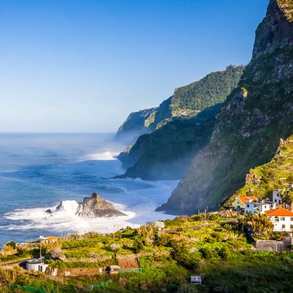 Weiße Häuser auf grünen Terrassenhängen unter steilen Klippen auf Madeira. Wellen brechen an Felsen entlang der blauen Atlantikküste.