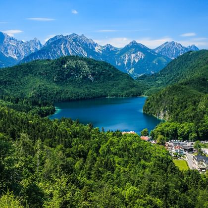 Luftaufnahme des Alpsees umgeben von bewaldeten Hügeln, Dorf Hohenschwangau mit Schloss und schneebedeckten Alpengipfeln unter blauem Himmel.